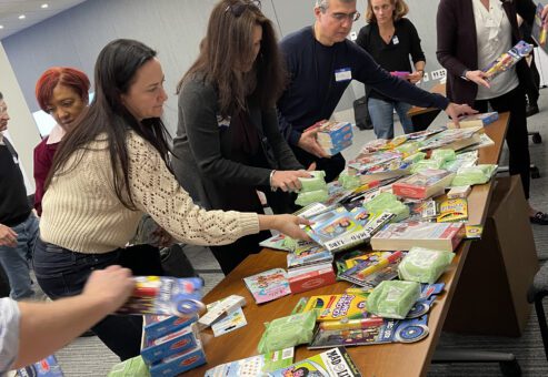 A group of individuals gather around a table filled with various school supplies, including pencils, erasers, and notebooks. They are sorting and organizing the supplies, as part of a team building CSR activity aimed at creating care packages for cancer patients.