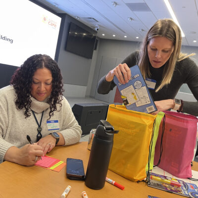 Two women participate in a team building activity as part of the 'Operation Cancer Care' event. One woman is writing a heartfelt note on colorful post-its, while the other is carefully packing a drawstring bag with puzzle books and supplies. Various materials, such as markers, a phone, and a water bottle, are spread across the table. A large screen in the background displays 'Team Building' and 'Operation Cancer Care,' adding context to the charitable activity. thumbnail