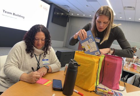 Two women participate in a team building activity as part of the 'Operation Cancer Care' event. One woman is writing a heartfelt note on colorful post-its, while the other is carefully packing a drawstring bag with puzzle books and supplies. Various materials, such as markers, a phone, and a water bottle, are spread across the table. A large screen in the background displays 'Team Building' and 'Operation Cancer Care,' adding context to the charitable activity.