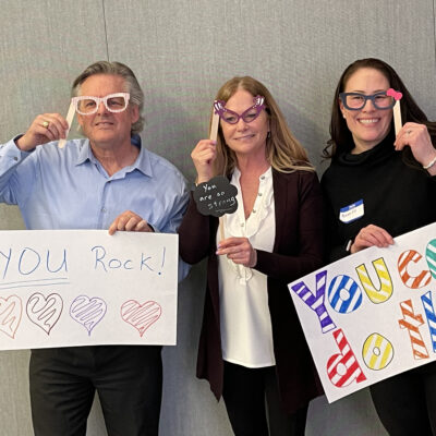 Three individuals holding up handmade motivational signs and wearing playful glasses during a team building event. The signs say 'YOU Rock!' with colorful hearts and 'You can do this,' encouraging messages meant to uplift and inspire. thumbnail