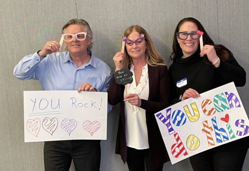 Three individuals holding up handmade motivational signs and wearing playful glasses during a team building event. The signs say 'YOU Rock!' with colorful hearts and 'You can do this,' encouraging messages meant to uplift and inspire.