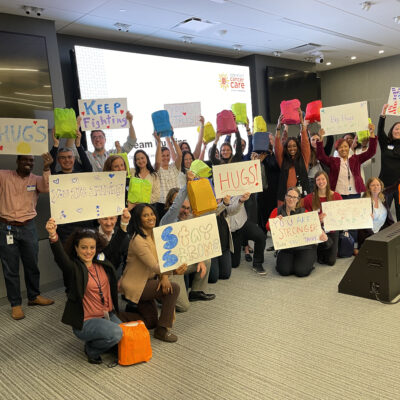 A group of people standing and kneeling in a conference room, smiling and holding up colorful posters and bags. The posters feature positive and supportive messages like 'Stay Strong,' 'HUGS,' 'You are Stronger than you Think,' and 'Keep Fighting.' Behind them is a presentation screen with the logo for 'Operation Cancer Care,' indicating this is part of a CSR team building event. thumbnail