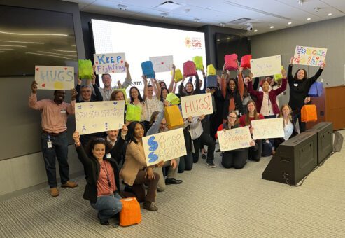A group of people standing and kneeling in a conference room, smiling and holding up colorful posters and bags. The posters feature positive and supportive messages like 'Stay Strong,' 'HUGS,' 'You are Stronger than you Think,' and 'Keep Fighting.' Behind them is a presentation screen with the logo for 'Operation Cancer Care,' indicating this is part of a CSR team building event.