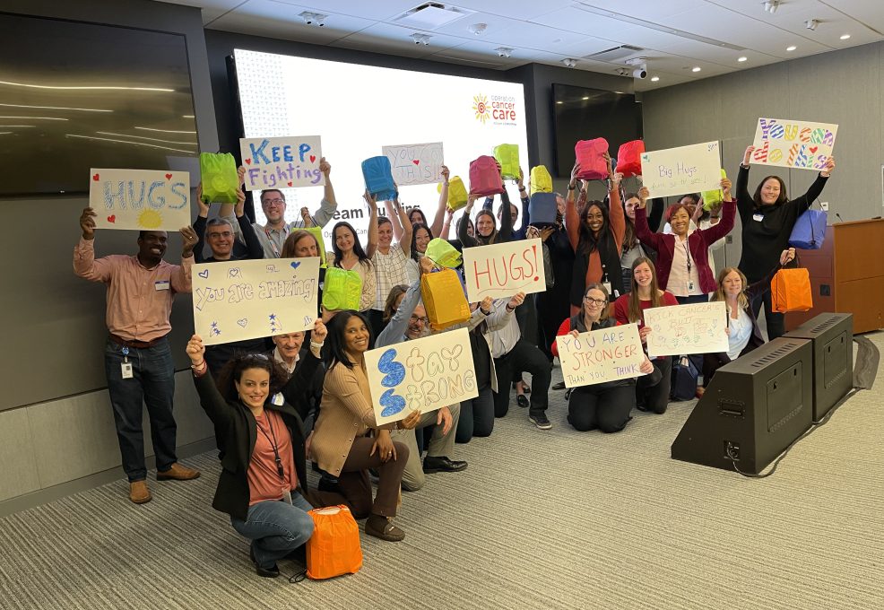 A group of people standing and kneeling in a conference room, smiling and holding up colorful posters and bags. The posters feature positive and supportive messages like 'Stay Strong,' 'HUGS,' 'You are Stronger than you Think,' and 'Keep Fighting.' Behind them is a presentation screen with the logo for 'Operation Cancer Care,' indicating this is part of a CSR team building event.