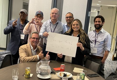 A group of seven people standing and seated around a table during a team building event. One woman holds a white sign that reads, 'You got this! You will be ok!' Others are smiling and giving thumbs up, showing supportive gestures.