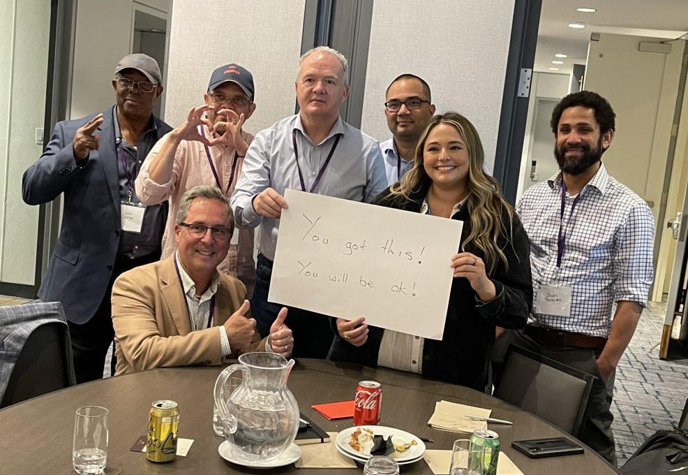 A group of seven people standing and seated around a table during a team building event. One woman holds a white sign that reads, 'You got this! You will be ok!' Others are smiling and giving thumbs up, showing supportive gestures.