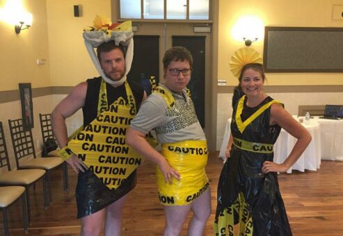 Three individuals pose confidently at a team building event, showcasing their creative outfits made from 'Caution' tape and other recyclable materials. The person on the left is wearing a black and yellow outfit, with a decorative headpiece made from caution tape. The person in the middle is also adorned in a yellow caution-tape skirt and a gray top. The individual on the right is dressed in a black and yellow dress with a matching headpiece, all made from caution tape and other materials. The mood is playful, and the group looks proud of their fashion designs.