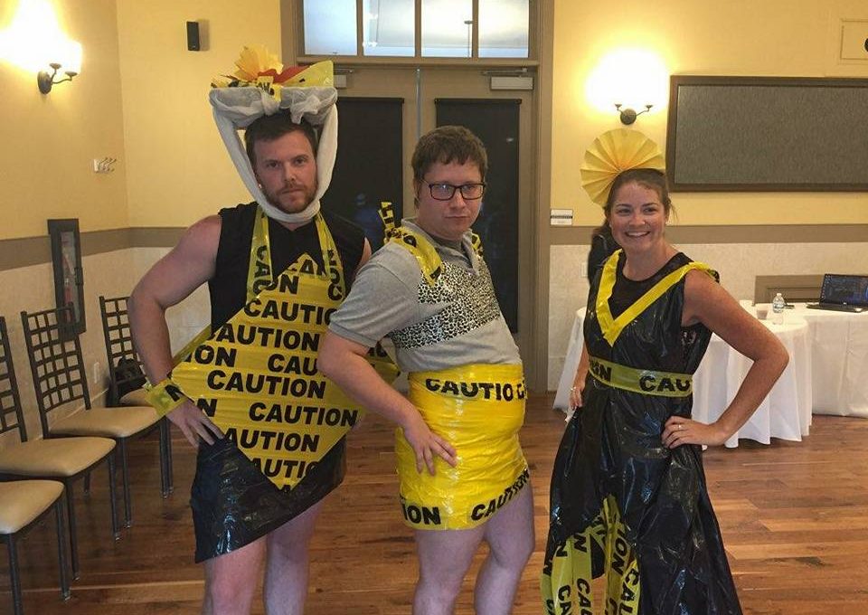 Three individuals pose confidently at a team building event, showcasing their creative outfits made from 'Caution' tape and other recyclable materials. The person on the left is wearing a black and yellow outfit, with a decorative headpiece made from caution tape. The person in the middle is also adorned in a yellow caution-tape skirt and a gray top. The individual on the right is dressed in a black and yellow dress with a matching headpiece, all made from caution tape and other materials. The mood is playful, and the group looks proud of their fashion designs.