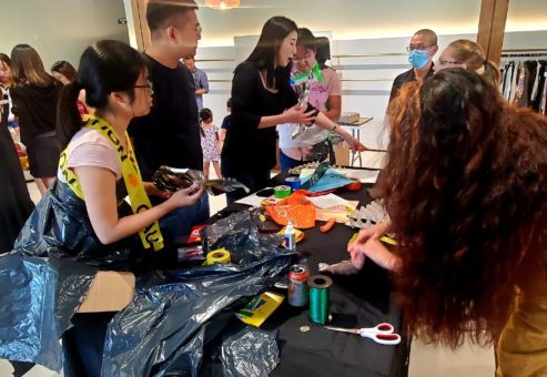 A group of participants engages in a Corporate Project Runway team building activity centered around creating fashion items. Several people are gathered around a table filled with various crafting materials such as scissors, ribbons, and recyclable items. Some participants are cutting and arranging materials while others are brainstorming or collaborating. One person wears an outfit made of trash bags and caution tape, reflecting the creative and playful nature of the event.