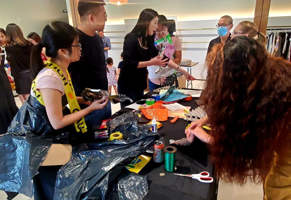 A group of participants engages in a Corporate Project Runway team building activity centered around creating fashion items. Several people are gathered around a table filled with various crafting materials such as scissors, ribbons, and recyclable items. Some participants are cutting and arranging materials while others are brainstorming or collaborating. One person wears an outfit made of trash bags and caution tape, reflecting the creative and playful nature of the event.