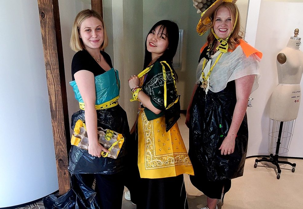 Three women standing together, smiling and posing for a photo during a team building activity. They are dressed in creative outfits made from various unconventional materials like trash bags, plastic, caution tape, and fabric. One of the women wears a large decorative hat made from a mix of recyclable materials.