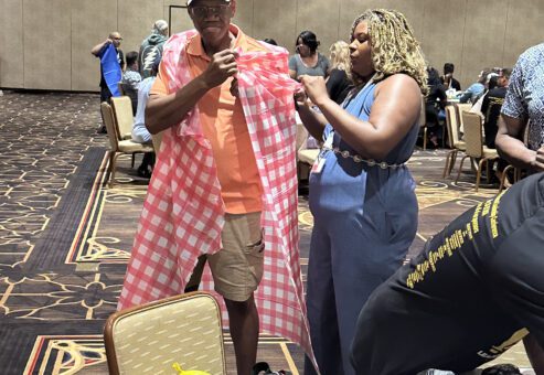 A man wearing a baseball cap is being fitted with a checkered red and white fabric draped over his shoulders, resembling a makeshift outfit. He is being assisted by a woman standing next to him, who is holding part of the fabric as she adjusts the look. They are participating in a fun team building activity in a large conference room with other groups in the background working on similar projects.