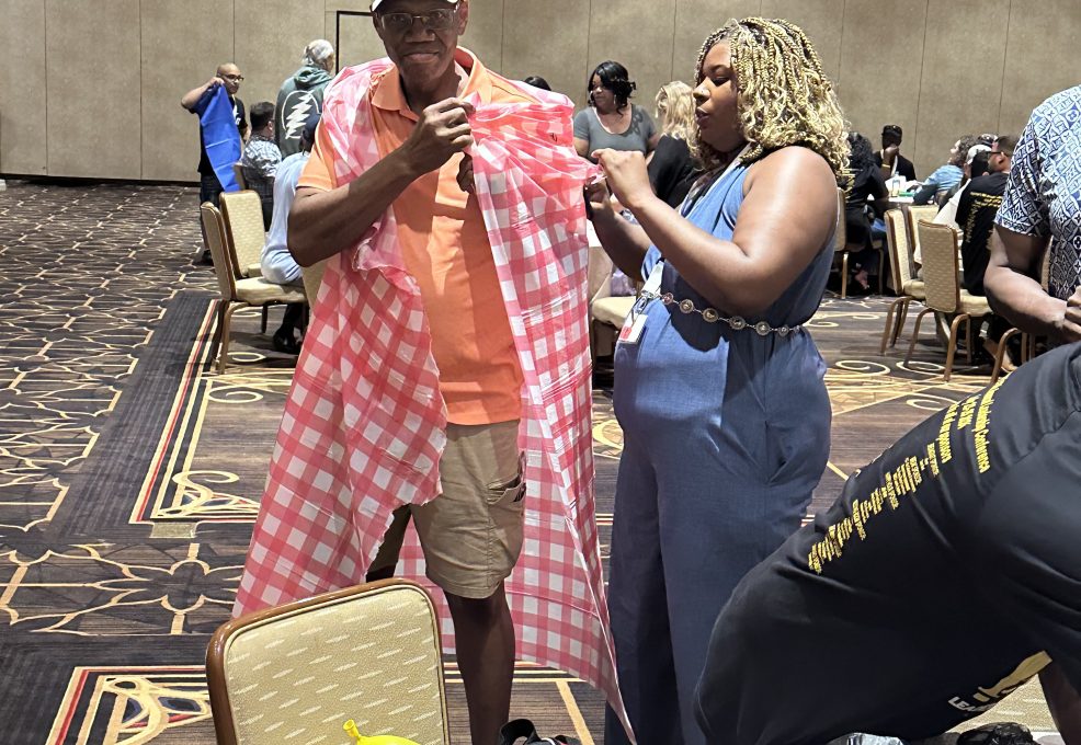 A man wearing a baseball cap is being fitted with a checkered red and white fabric draped over his shoulders, resembling a makeshift outfit. He is being assisted by a woman standing next to him, who is holding part of the fabric as she adjusts the look. They are participating in a fun team building activity in a large conference room with other groups in the background working on similar projects.