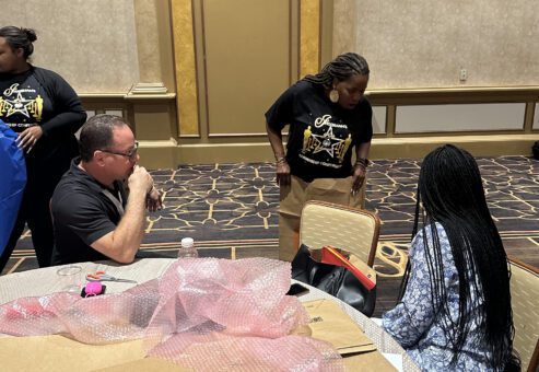 A group of people seated around a table in a large conference room working on a team building project. The table is covered with various crafting supplies, including bubble wrap, cardboard, markers, and a sketchpad. One man is seated, appearing to work or think while another person, seated next to him, is writing on paper. A woman in a black t-shirt is standing near the table, holding a piece of material or cardboard involved in the activity. The participants are engaging in a creative task, designing a garment together.