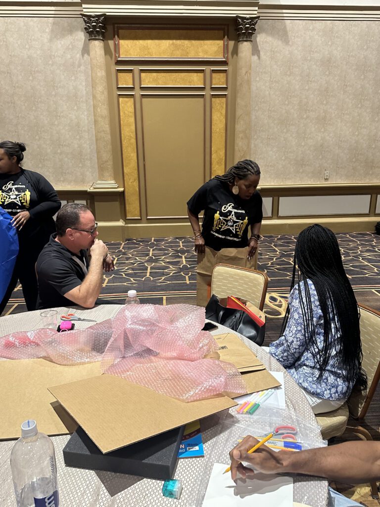 A group of people seated around a table in a large conference room working on a team building project. The table is covered with various crafting supplies, including bubble wrap, cardboard, markers, and a sketchpad. One man is seated, appearing to work or think while another person, seated next to him, is writing on paper. A woman in a black t-shirt is standing near the table, holding a piece of material or cardboard involved in the activity. The participants are engaging in a creative task, designing a garment together.
