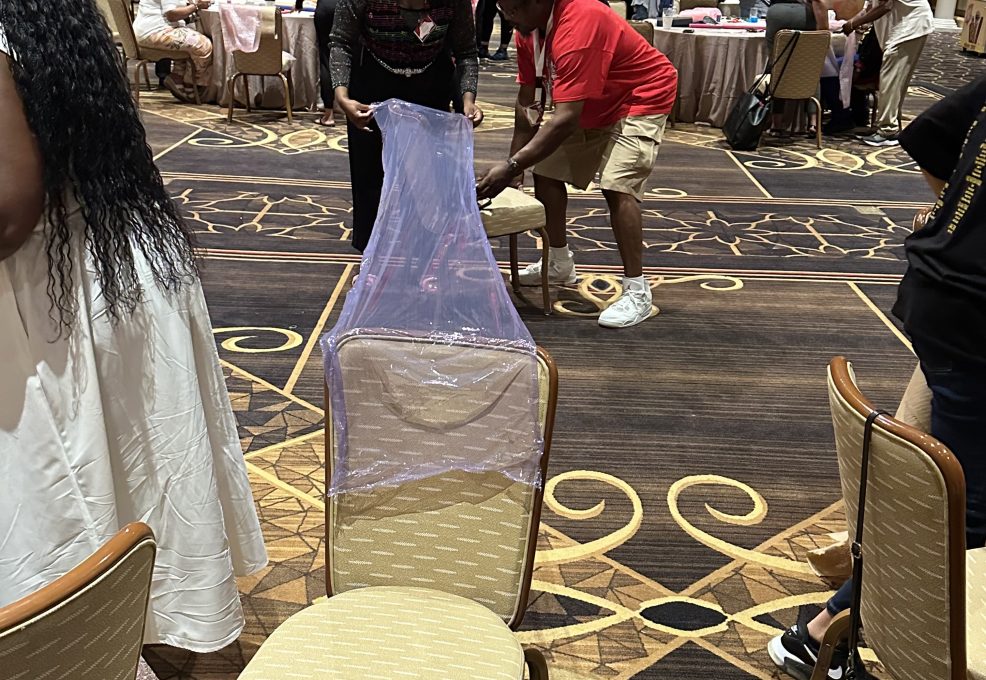 Two participants in a team building activity are working together to cover a chair with a transparent purple material. The man is securing the fabric at the bottom, while the woman stretches the material across the back of the chair.