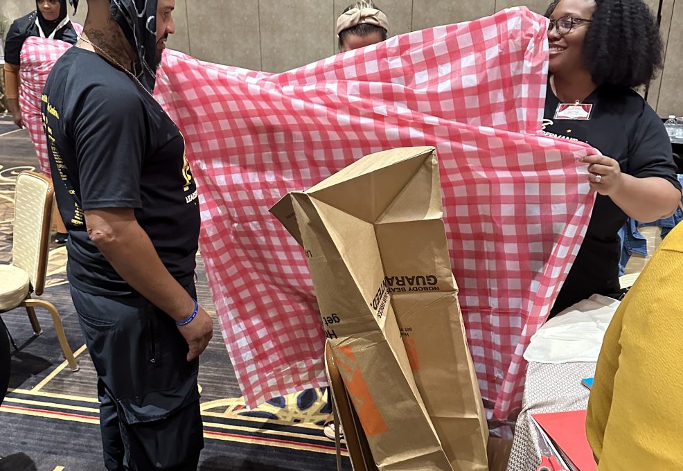 Two participants at a team building event stand, with one holding a large red and white checkered fabric, while the other appears to be modeling it or assisting in its setup. Cardboard and other recyclable materials are visible, being used to create an outfit.