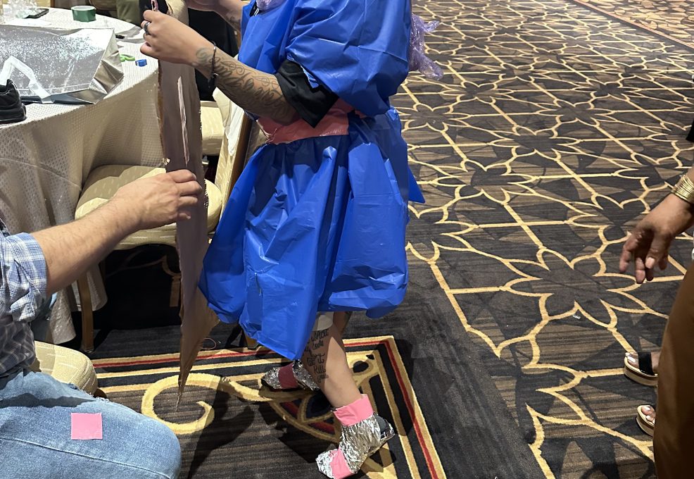 A smiling participant wearing a creatively made outfit composed of colorful recyclable materials, standing in a ballroom. The person is holding a piece of material while others in the background work on their designs. It is a team building event themed around fashion design, with participants using unconventional materials to create outfits.