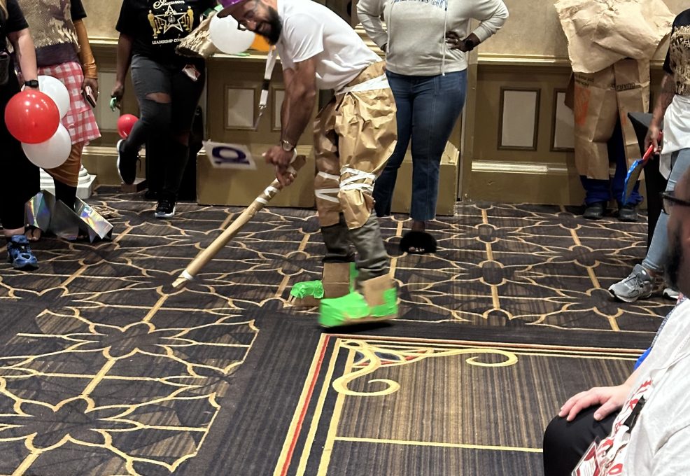 A group of people participate in a fun team building event inspired by a fashion runway show. The focus is on one individual wearing a creative outfit made from paper and other recyclable materials, including a purple hat, brown pants, and neon green shoes. The individual is in an action pose, holding a stick as if preparing for a playful activity. Several other participants, also dressed in quirky, recyclable material outfits, stand around watching.