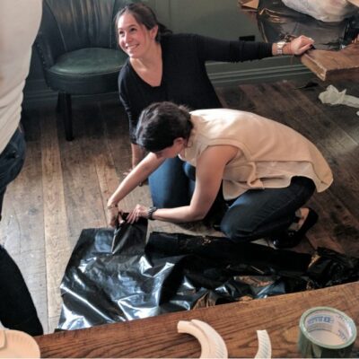 Two women engaged in a crafting activity for a team building event. One is kneeling on the wooden floor, cutting or adjusting a piece of black plastic material, while the other woman leans on a nearby table, smiling and watching. thumbnail