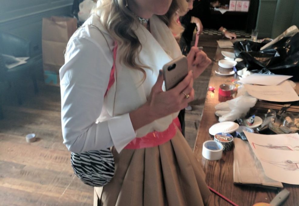 A woman standing near a table full of crafting materials, wearing a creative outfit made of paper or fabric for a team building event. Another participant is seen working in the background, focused on the materials laid out.