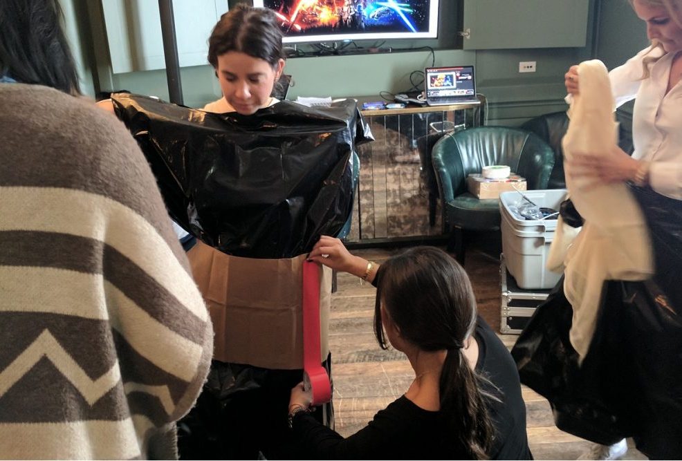 Participants collaborate during a Corporate Project Runway team building fashion activity. A woman stands in the center while others assist in constructing a garment out of black plastic material and cardboard. One person is adjusting the fit by attaching materials around her waist.