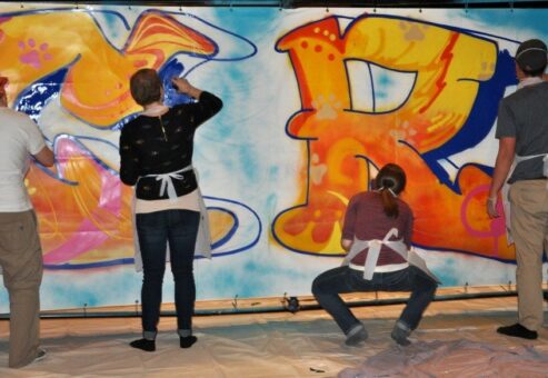 Participants wearing aprons work together to paint a colorful graffiti mural on a large canvas during a Custom Team Building event.