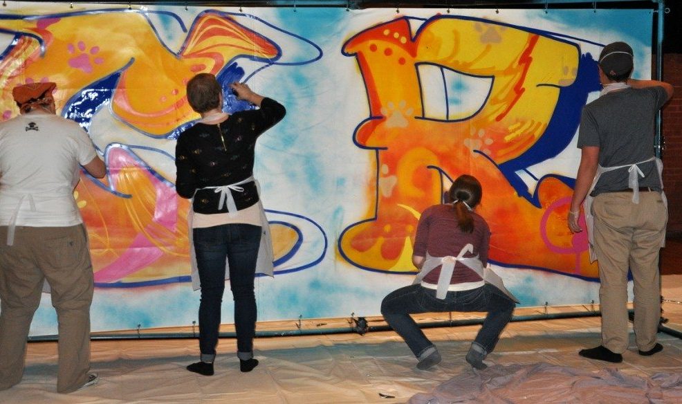 Participants wearing aprons work together to paint a colorful graffiti mural on a large canvas during a Custom Team Building event.