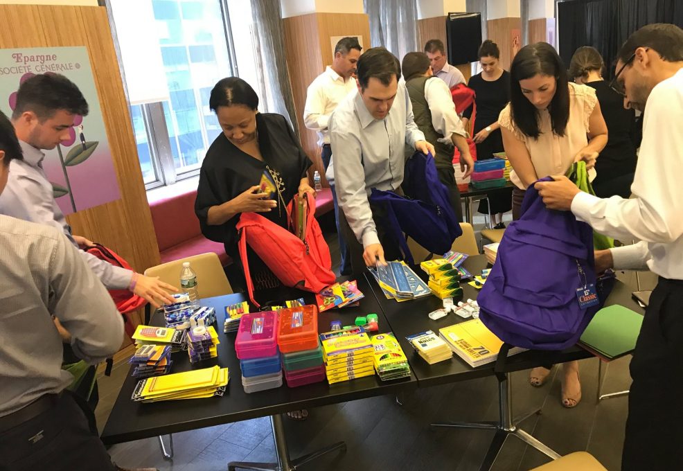 Corporate team members packing backpacks with school supplies during a csr team building event. The table is filled with notebooks, pencils, and other educational materials as participants work together to prepare donations, emphasizing teamwork and giving back to the community.