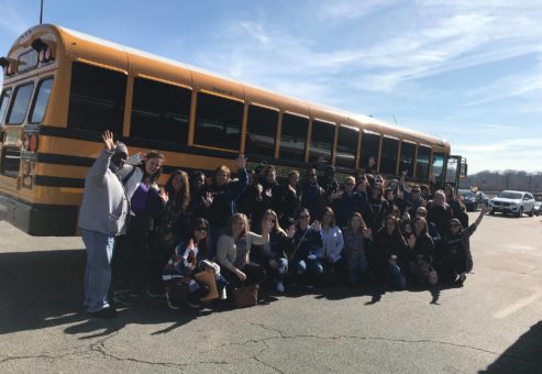 A large group of people poses in front of a yellow school bus on a sunny day, many of them waving at the camera. They are outdoors in a parking lot, with clear skies above, creating a sense of enthusiasm and camaraderie. The group appears to be preparing for or returning from a volunteer outing, part of a team building event. The mix of smiles and waves highlights the positive energy and collaborative spirit of the group.