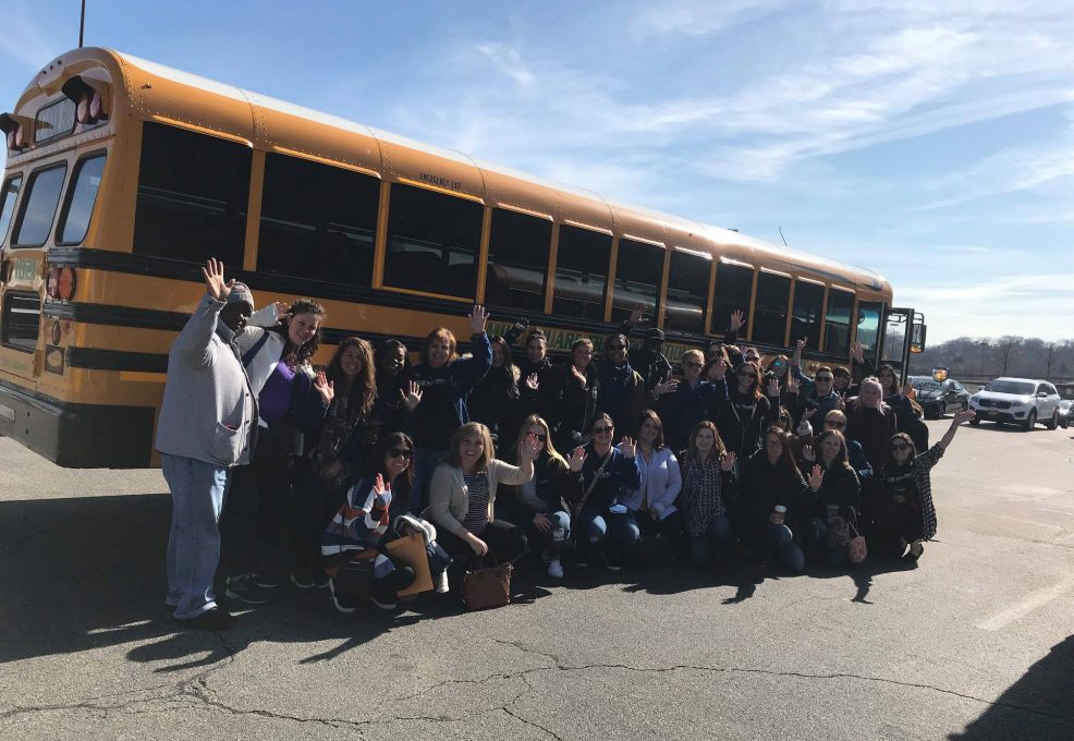 A large group of people poses in front of a yellow school bus on a sunny day, many of them waving at the camera. They are outdoors in a parking lot, with clear skies above, creating a sense of enthusiasm and camaraderie. The group appears to be preparing for or returning from a volunteer outing, part of a team building event. The mix of smiles and waves highlights the positive energy and collaborative spirit of the group.
