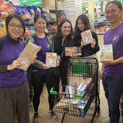 A group of five volunteers stands together in a grocery store aisle, smiling and holding up various food items. They have a shopping cart filled with packaged goods for a donation or charity event. They are participating in a Do Good Bus team building event where they are collecting items to support a community cause. The atmosphere appears cheerful and collaborative. thumbnail