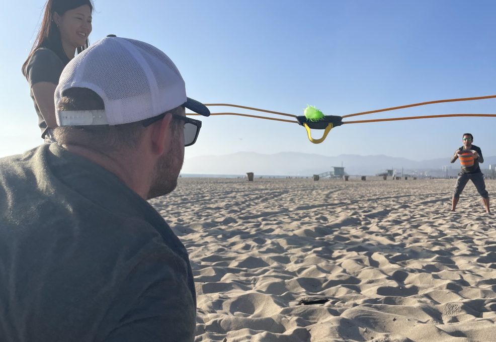 Team members use a slingshot during a Corporate Survivor beach activity, focusing on strategy and team coordination.