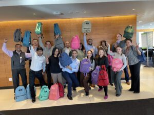 A group of smiling participants proudly holding up backpacks filled with school supplies during a Tools for Schools CSR team building event.