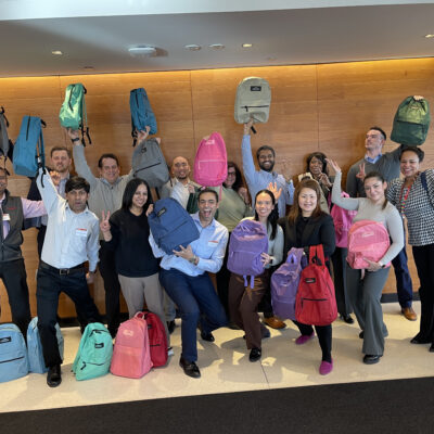 A group of smiling participants proudly holding up backpacks filled with school supplies during a Tools for Schools CSR team building event.