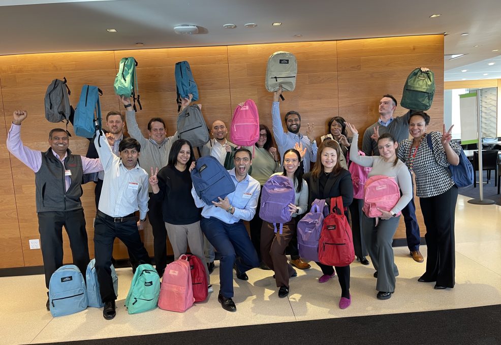 A group of smiling participants proudly holding up backpacks filled with school supplies during a Tools for Schools CSR team building event.