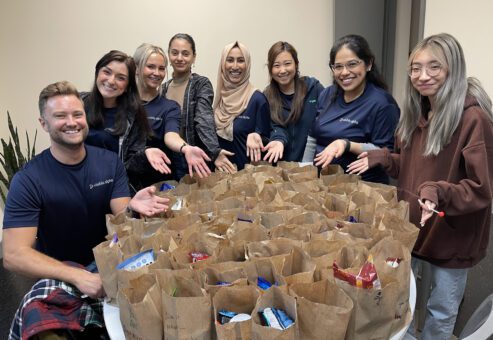A group of eight smiling individuals stand around a table filled with brown paper bags, likely packed with food or care items. They are proudly showing off their work, extending their hands toward the bags. Everyone in the group appears happy and engaged in this Do Good Bus team building event.