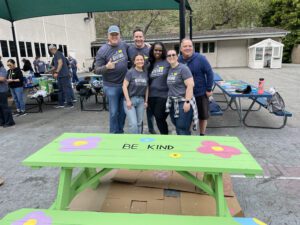 A group of six people standing together, smiling in front of a brightly painted picnic table that reads "BE KIND" in the center. The picnic table is painted green with colorful flowers. All participants are wearing matching gray "Giving" shirts, indicating they are part of a charitable team building event. The surrounding area shows other tables and people engaged in similar activities, painting or working on projects.