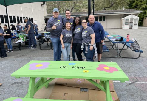 A group of six people standing together, smiling in front of a brightly painted picnic table that reads 
