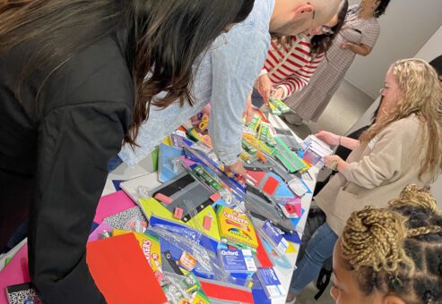 Participants at a Tools for Schools CSR event gather around a table filled with school supplies as they pack colorful folders, notebooks, and other materials into backpacks to give to students.