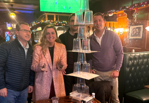 A group of team members proudly displaying their impressive tower of stacked cups during the Pub Crawl Scavenger Hunt, showcasing creativity and teamwork in a lively bar setting.