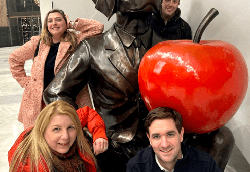Team posing with a playful dog statue and bright red apple during the Pub Crawl Scavenger Hunt, blending exploration and friendly competition for a memorable team building experience.
