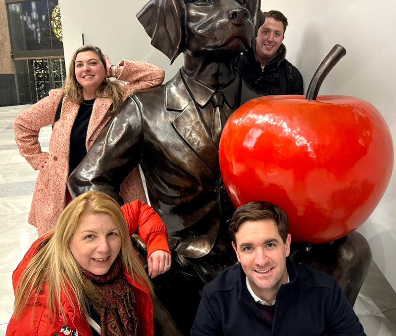 Team posing with a playful dog statue and bright red apple during the Pub Crawl Scavenger Hunt, blending exploration and friendly competition for a memorable team building experience.