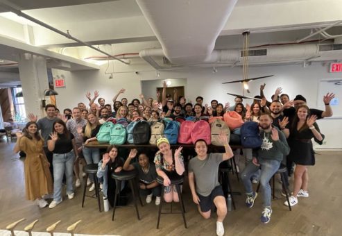 Large group of smiling participants posing with colorful backpacks filled with school supplies after a successful corporate csr event, highlighting teamwork and community impact through the Tools for Schools program.