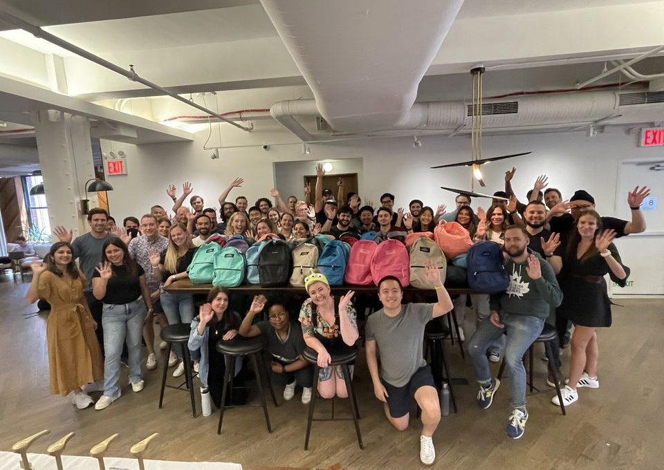 Large group of smiling participants posing with colorful backpacks filled with school supplies after a successful corporate csr event, highlighting teamwork and community impact through the Tools for Schools program.