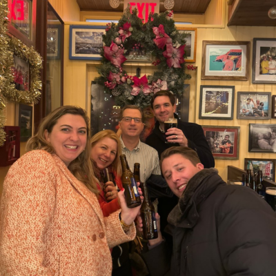 Smiling team members raise their drinks in celebration during a festive stop on the Pub Crawl Scavenger Hunt, enjoying the holiday decor and camaraderie. thumbnail