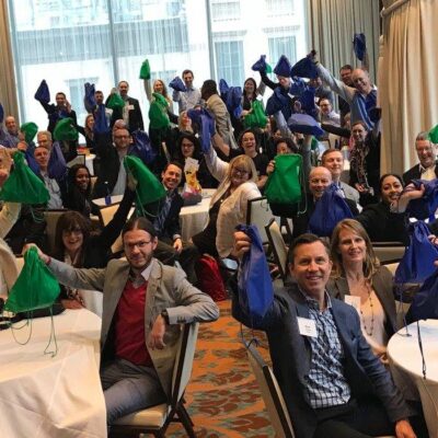 A large group of participants sits at round tables, proudly holding up green and blue drawstring bags filled with care kits during the Helping Hands team building event. The room is filled with energy as the group celebrates their collective effort to help the community. thumbnail