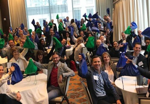 A large group of participants sits at round tables, proudly holding up green and blue drawstring bags filled with care kits during the Helping Hands team building event. The room is filled with energy as the group celebrates their collective effort to help the community.