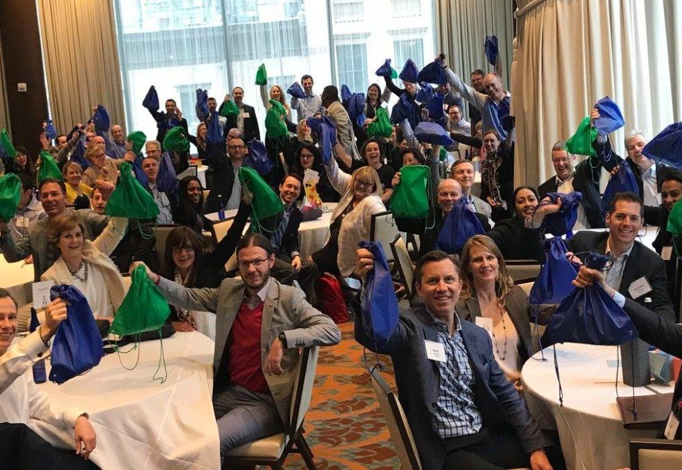 A large group of participants sits at round tables, proudly holding up green and blue drawstring bags filled with care kits during the Helping Hands team building event. The room is filled with energy as the group celebrates their collective effort to help the community.