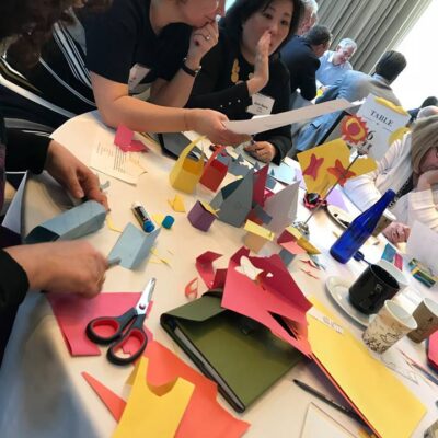 Participants work together on a creative task involving colorful paper cutouts during the Helping Hands team building event. They collaborate and discuss their progress while assembling materials for care kits.